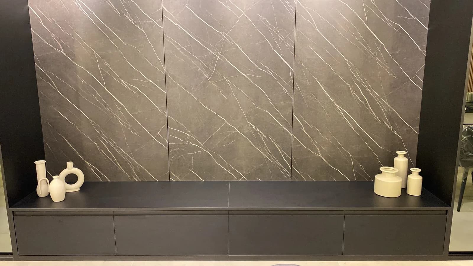Aparador baixo preto com vasos claros diante de painel marmorizado grafite
