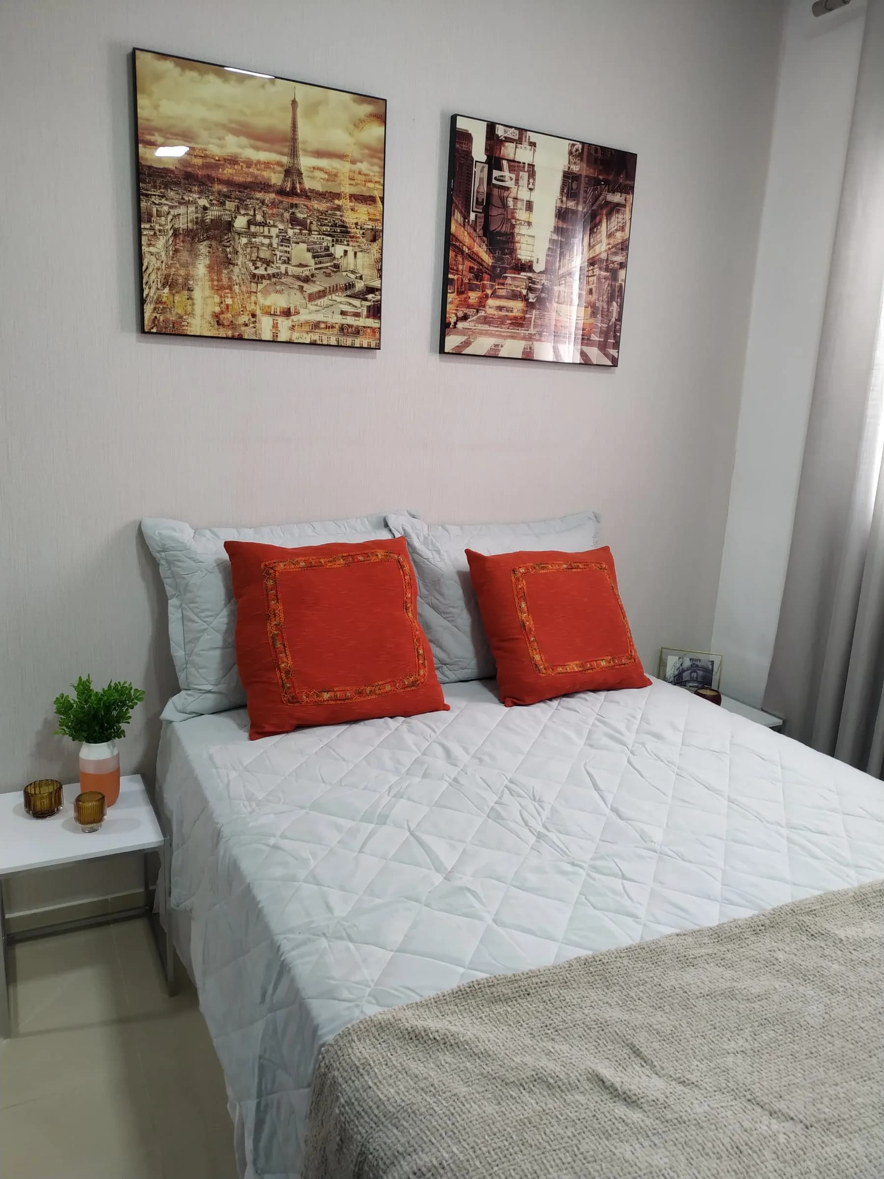 Quarto de casal com roupa de cama branca, almofadas laranja e dois quadros urbanos